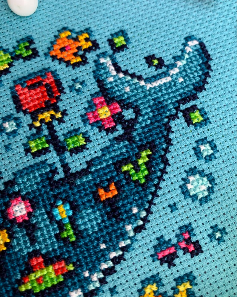 Puede incluir: Primer plano de un dise&ntilde;o de punto de cruz sobre tela azul claro. El dise&ntilde;o presenta una forma azul grande y estilizada con acentos blancos, rodeada de elementos florales y geom&eacute;tricos coloridos en rojo, naranja, verde y rosa. Las puntadas son claramente visibles.
