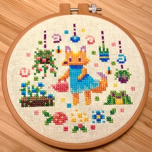 May include: A cross-stitch embroidery hoop depicting a fox in a blue dress watering a garden with flowers, butterflies, and other garden elements.