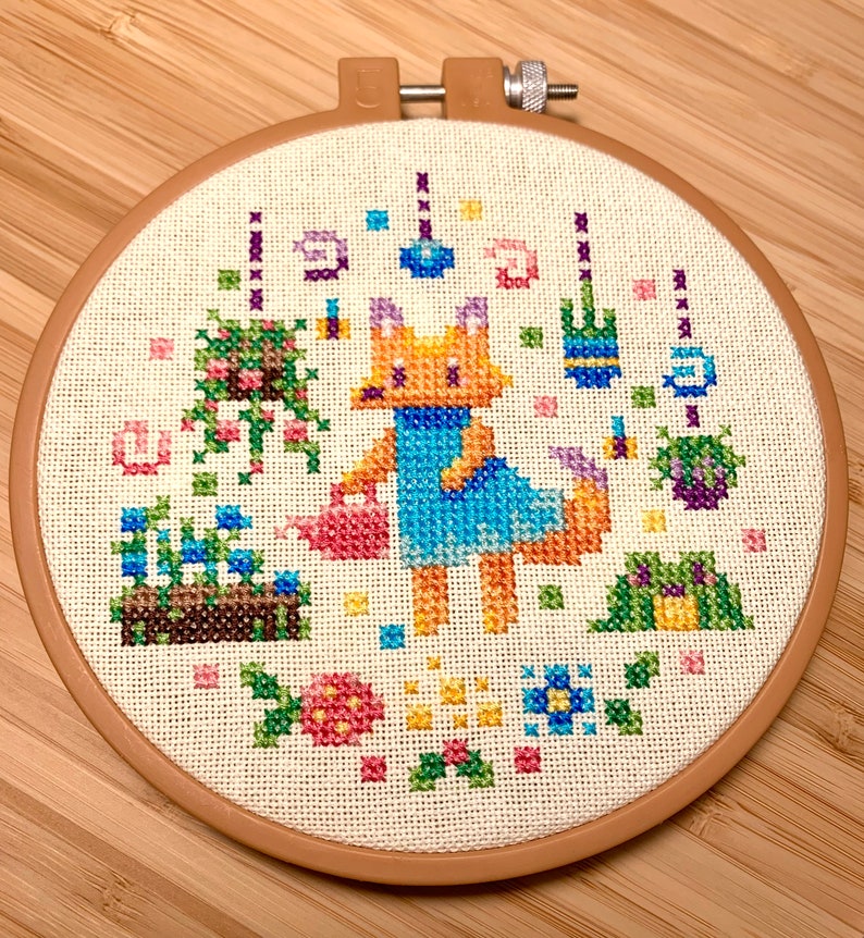 May include: A cross-stitch embroidery hoop depicting a fox in a blue dress watering a garden with flowers, butterflies, and other garden elements.