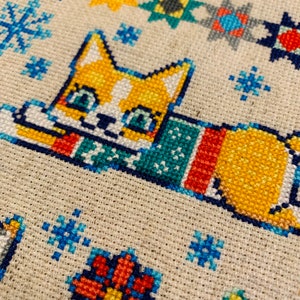 Peut inclure: Un motif de point de croix repr&eacute;sentant un chat portant une &eacute;charpe color&eacute;e, entour&eacute; de flocons de neige et d'autres formes g&eacute;om&eacute;triques. Le chat est orange et blanc avec des yeux bleus.