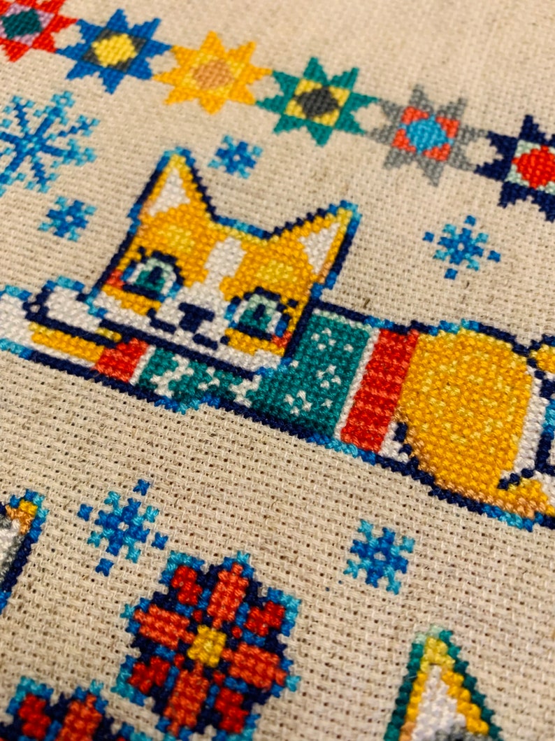 Peut inclure: Un motif de point de croix repr&eacute;sentant un chat portant une &eacute;charpe color&eacute;e, entour&eacute; de flocons de neige et d'autres formes g&eacute;om&eacute;triques. Le chat est orange et blanc avec des yeux bleus.