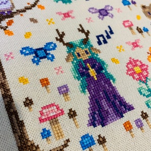 May include: Cross-stitch embroidery featuring a purple-clad fairy with antlers, surrounded by mushrooms, butterflies, and pumpkins. The fairy has green hair and a yellow crown.