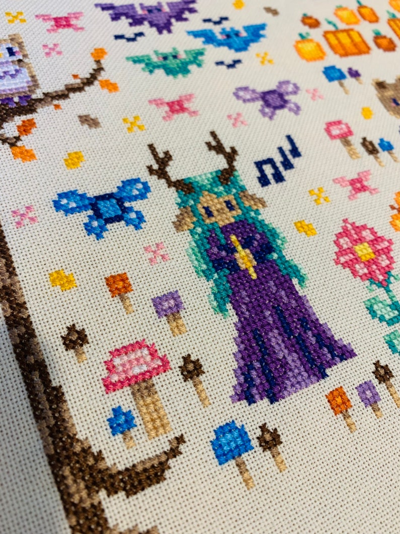 May include: Cross-stitch embroidery featuring a purple-clad fairy with antlers, surrounded by mushrooms, butterflies, and pumpkins. The fairy has green hair and a yellow crown.