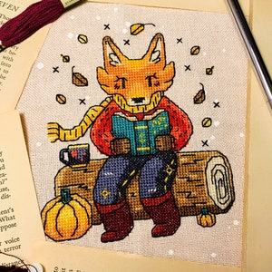 May include: A cross-stitch pattern of a fox wearing a red sweater and a yellow scarf, sitting on a log with a book in its paws. The fox is surrounded by autumn leaves and a pumpkin.