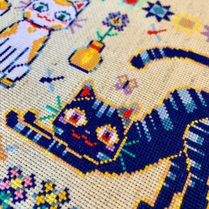 May include: Cross-stitch embroidery featuring two cats, one white with orange markings and the other black with blue stripes, surrounded by colorful flowers and geometric patterns.