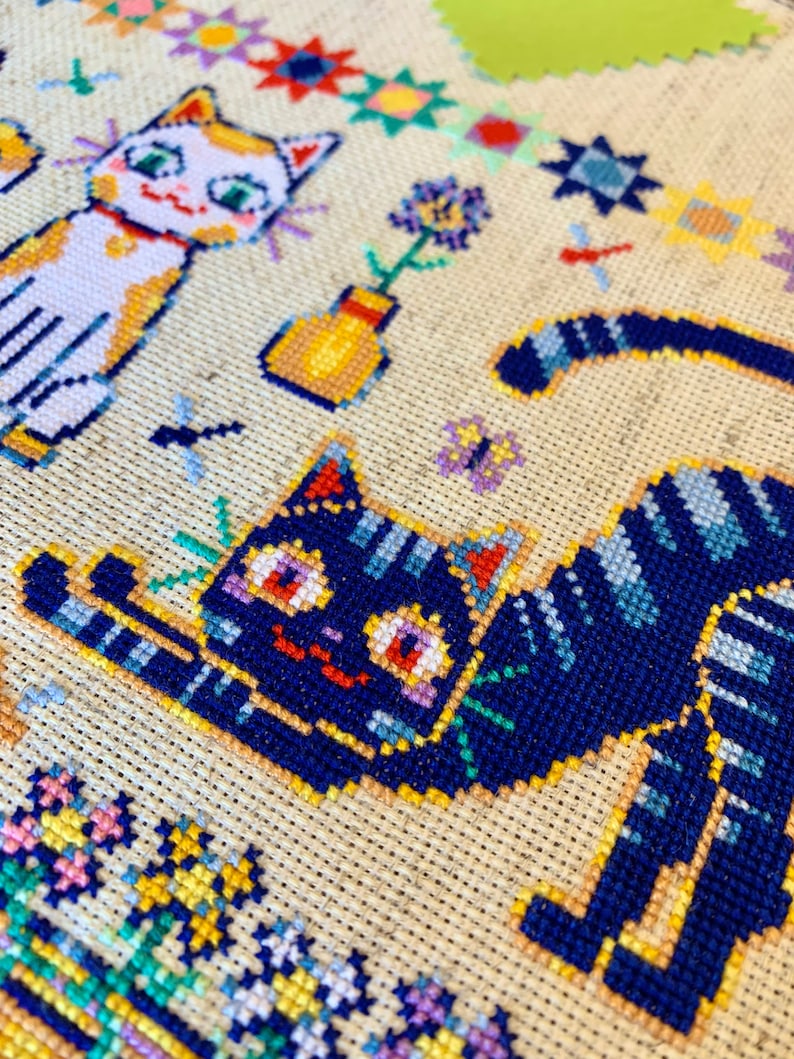 May include: Cross-stitch embroidery featuring two cats, one white with orange markings and the other black with blue stripes, surrounded by colorful flowers and geometric patterns.