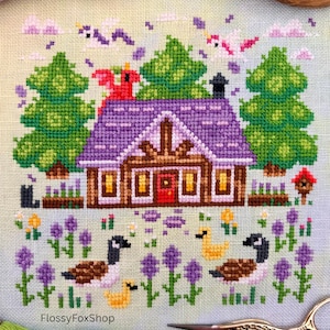 Op de afbeelding: Kruissteekontwerp met een hut met een paars dak, groene bomen en ganzen. De sc&egrave;ne bevat paarse bloemen, wolken en vogels. De tekst "FlossyFoxShop" is zichtbaar. Borduurgaren en schaar op de voorgrond.