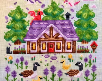 May include: Cross-stitch design featuring a cabin with a purple roof, green trees, and geese. The scene includes purple flowers, clouds, and birds. The text "FlossyFoxShop" is visible. Embroidery floss and scissors are in the foreground.