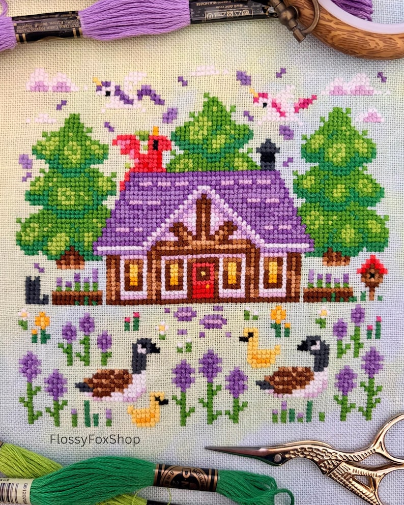 Op de afbeelding: Kruissteekontwerp met een hut met een paars dak, groene bomen en ganzen. De sc&egrave;ne bevat paarse bloemen, wolken en vogels. De tekst "FlossyFoxShop" is zichtbaar. Borduurgaren en schaar op de voorgrond.