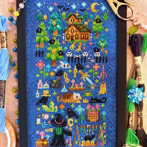 May include: A framed cross-stitch embroidery on blue fabric featuring a fantasy scene with a house, trees, a crescent moon, stars, a sword, a cauldron, a cat, and a figure in a hat. The text FlossyFoxShop is printed in the bottom right corner.