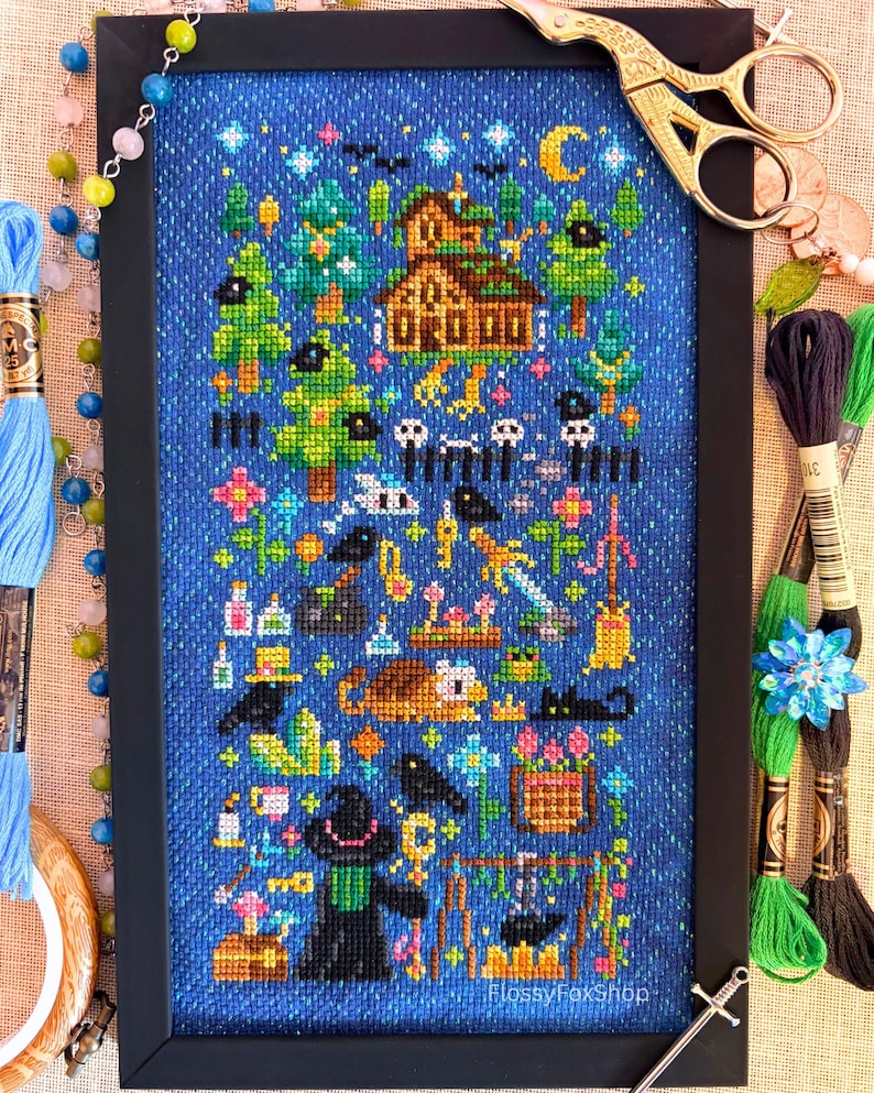 May include: A framed cross-stitch embroidery on blue fabric featuring a fantasy scene with a house, trees, a crescent moon, stars, a sword, a cauldron, a cat, and a figure in a hat. The text FlossyFoxShop is printed in the bottom right corner.