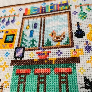 May include: Cross-stitch embroidery of a small house with a green roof, a brown door, and a window with a bookshelf above it. There are three stools at a counter inside the house.
