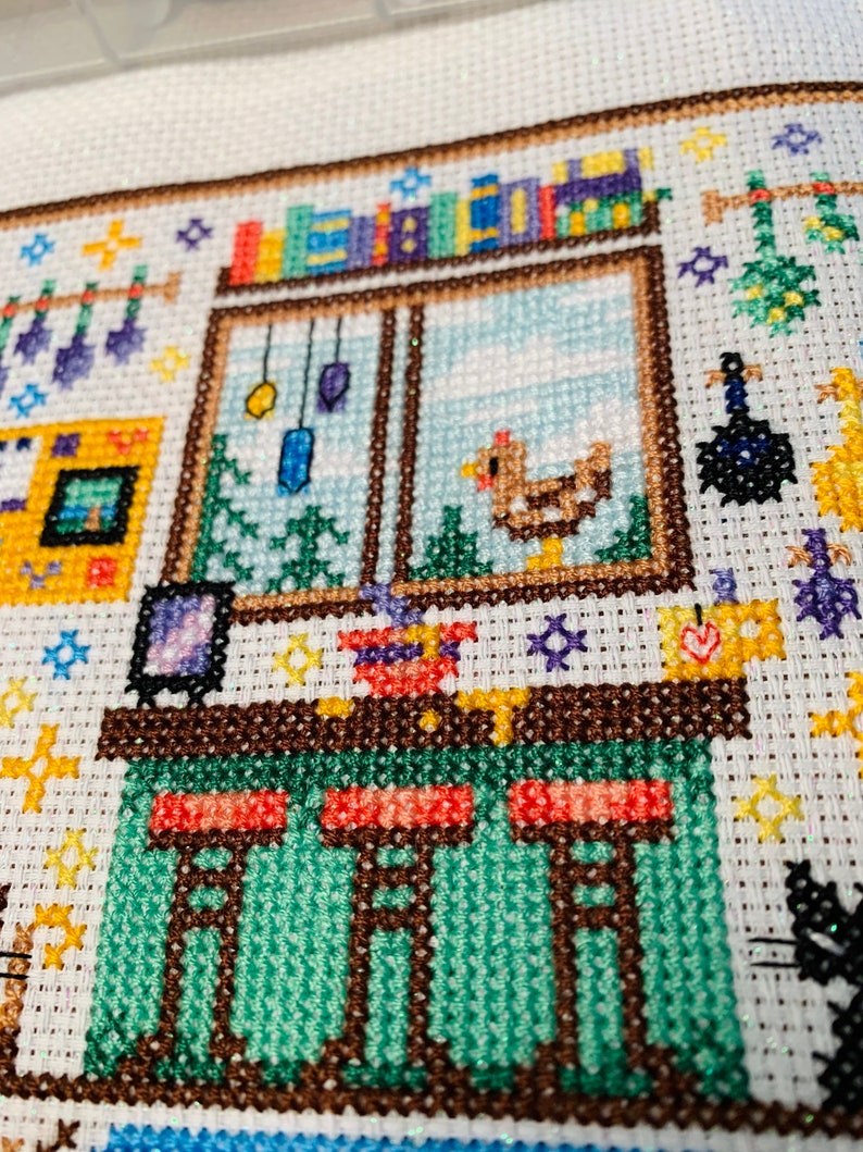 May include: Cross-stitch embroidery of a small house with a green roof, a brown door, and a window with a bookshelf above it. There are three stools at a counter inside the house.