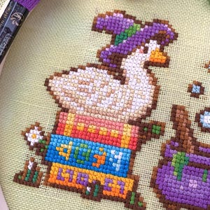 May include: Cross-stitch embroidery featuring a white goose in a purple hat, sitting on colourful books. The books are labelled with the word "LIBRA" in orange and blue. A small white flower and purple embroidery thread are also visible.