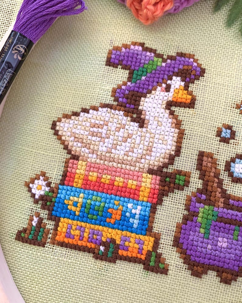 May include: Cross-stitch embroidery featuring a white goose in a purple hat, sitting on colourful books. The books are labelled with the word "LIBRA" in orange and blue. A small white flower and purple embroidery thread are also visible.