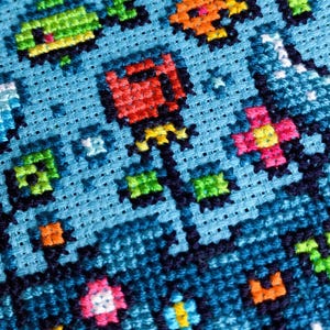Puede incluir: Primer plano de un dise&ntilde;o de punto de cruz sobre tela azul claro. El dise&ntilde;o presenta flores pixeladas coloridas y otras formas en tonos de rojo, naranja, rosa, verde y amarillo. Las puntadas est&aacute;n cuidadosamente alineadas.
