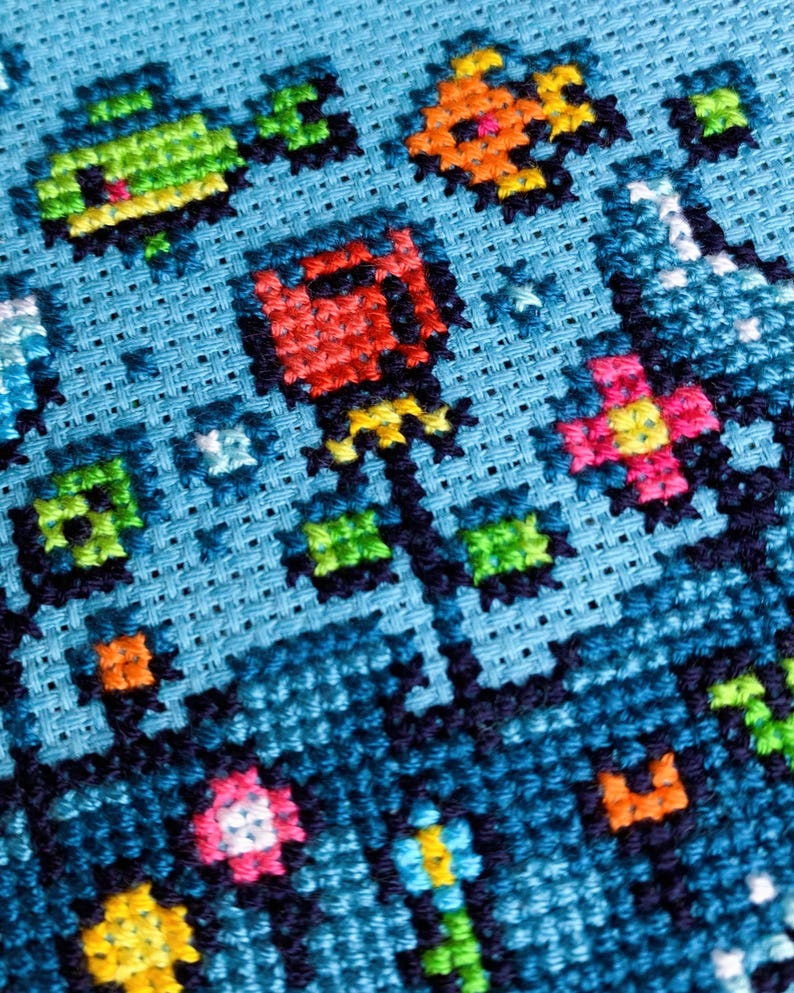Puede incluir: Primer plano de un dise&ntilde;o de punto de cruz sobre tela azul claro. El dise&ntilde;o presenta flores pixeladas coloridas y otras formas en tonos de rojo, naranja, rosa, verde y amarillo. Las puntadas est&aacute;n cuidadosamente alineadas.