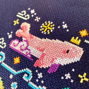 May include: A pink and purple cross-stitch design of a whale swimming in a blue ocean with yellow stars and a white snowflake.