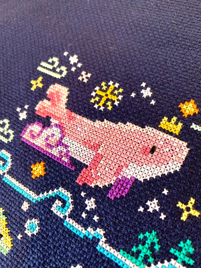 May include: A pink and purple cross-stitch design of a whale swimming in a blue ocean with yellow stars and a white snowflake.