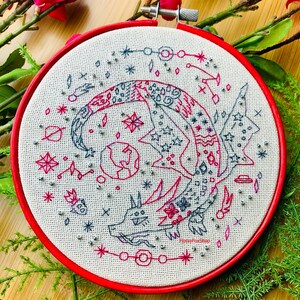 May include: Embroidered hoop art featuring a stylized dragon with a red, pink, and gray color scheme. The dragon is surrounded by stars, planets, and other celestial imagery. The hoop is red and the fabric is white.