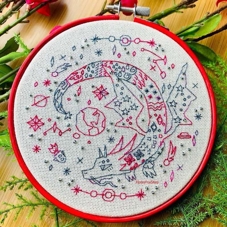 Peut inclure: Art de broderie en forme de cercle repr&eacute;sentant un dragon stylis&eacute; aux couleurs rouge, rose et gris. Le dragon est entour&eacute; d'&eacute;toiles, de plan&egrave;tes et d'autres images c&eacute;lestes. Le cercle est rouge et le tissu est blanc.
