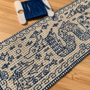 K&ouml;nnte beinhalten: Nahaufnahme eines blauen und wei&szlig;en Kreuzstich-Stickprojekts. Das Design zeigt einen stilisierten Vogel mit einem geometrischen Muster. Die Stickerei ist mit einem blauen Rand eingefasst.