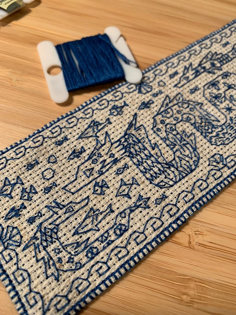 K&ouml;nnte beinhalten: Nahaufnahme eines blauen und wei&szlig;en Kreuzstich-Stickprojekts. Das Design zeigt einen stilisierten Vogel mit einem geometrischen Muster. Die Stickerei ist mit einem blauen Rand eingefasst.