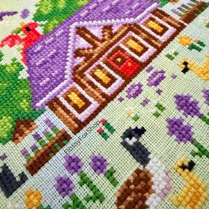 Op de afbeelding: Kruissteekborduurwerk met een gedetailleerde landelijke sc&egrave;ne. Een huis met een paars dak en bruine afwerking, met een rode deur, is omgeven door bloemen, vogels en ganzen. De tekst "FlossyFoxShop" is zichtbaar.