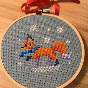 May include: A blue and white cross-stitch embroidery of a fox wearing a blue scarf and purple ice skates on a light blue background with white snowflakes.