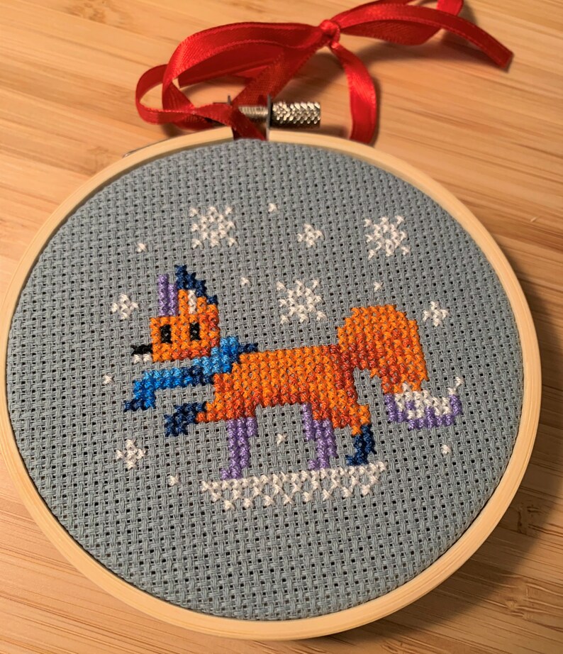 May include: A blue and white cross-stitch embroidery of a fox wearing a blue scarf and purple ice skates on a light blue background with white snowflakes.