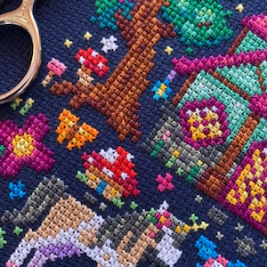 May include: A close-up of a cross-stitch embroidery on navy fabric. The design depicts a fantasy forest scene with a tree, mushrooms, flowers, and a unicorn. The colour scheme includes browns, greens, pinks, and purples.