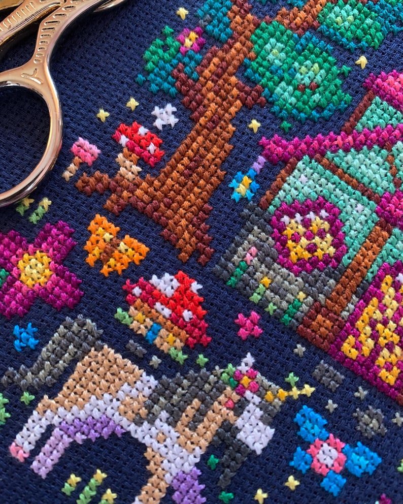 May include: A close-up of a cross-stitch embroidery on navy fabric. The design depicts a fantasy forest scene with a tree, mushrooms, flowers, and a unicorn. The colour scheme includes browns, greens, pinks, and purples.