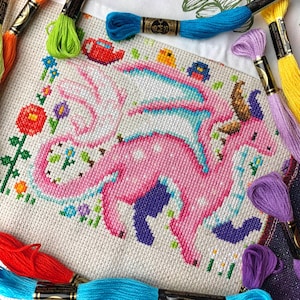 May include: A cross-stitch project featuring a pink dragon with blue and white accents, surrounded by colorful flowers and greenery. The dragon has large wings and is set against a white background. Embroidery floss in various colors, including orange, green, and purple, frames the artwork.