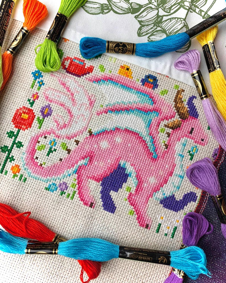 May include: A cross-stitch project featuring a pink dragon with blue and white accents, surrounded by colorful flowers and greenery. The dragon has large wings and is set against a white background. Embroidery floss in various colors, including orange, green, and purple, frames the artwork.