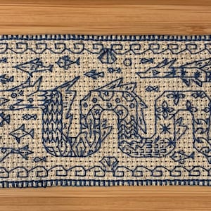 K&ouml;nnte beinhalten: Ein blaues und wei&szlig;es besticktes Stoffpanel, das eine stilisierte Seeschlange mit Fischen und anderen Meereslebewesen darstellt. Der Stoff ist mit einem strukturierten Muster gewebt.