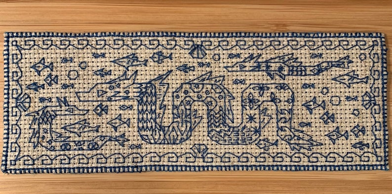 K&ouml;nnte beinhalten: Ein blaues und wei&szlig;es besticktes Stoffpanel, das eine stilisierte Seeschlange mit Fischen und anderen Meereslebewesen darstellt. Der Stoff ist mit einem strukturierten Muster gewebt.