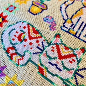 May include: Close-up of a cross-stitch embroidery featuring a white cat with red and green details. The cat is surrounded by other colorful designs, including flowers and a butterfly.