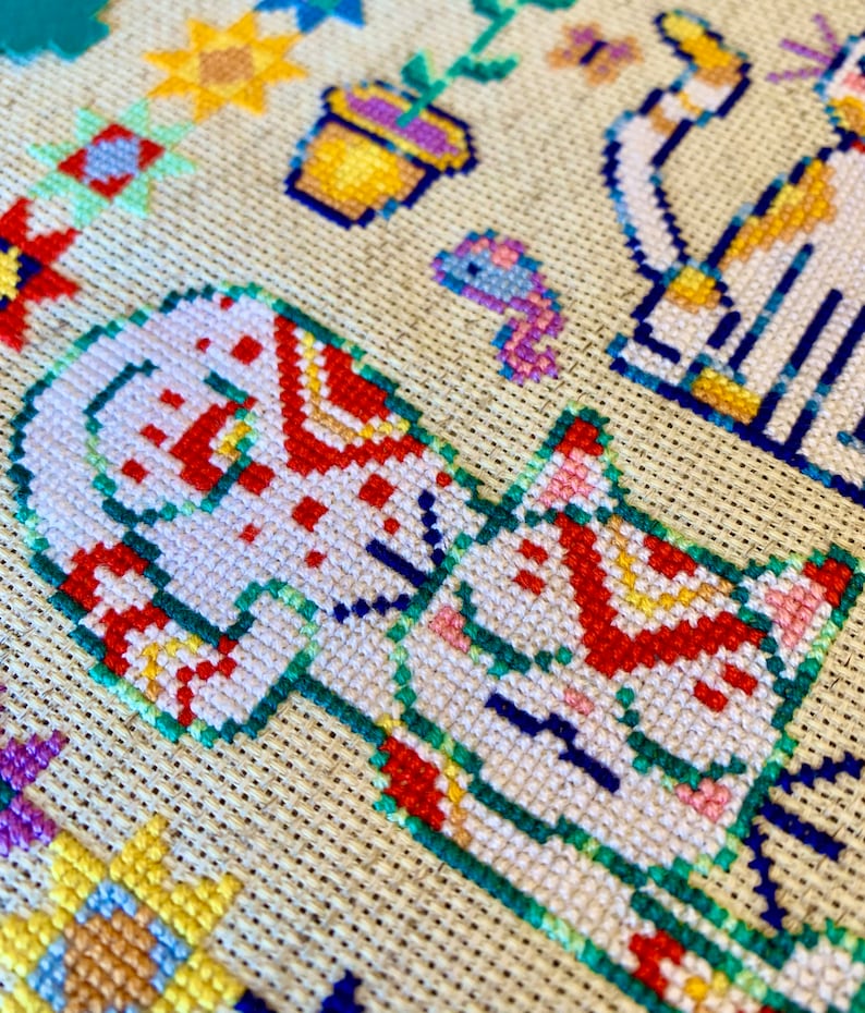 May include: Close-up of a cross-stitch embroidery featuring a white cat with red and green details. The cat is surrounded by other colorful designs, including flowers and a butterfly.