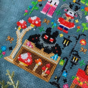 May include: Cross-stitch pattern featuring a black bat with red eyes, a red and white mushroom, and a small house with a brown roof and a yellow door. The pattern is on a blue background with other colorful details.