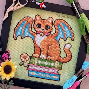 May include: Cross-stitch art of an orange cat with blue and orange wings, sitting on a stack of books. The artwork is framed in black and surrounded by embroidery floss, scissors, and a charm. The text "FlossyFoxShop" is visible.