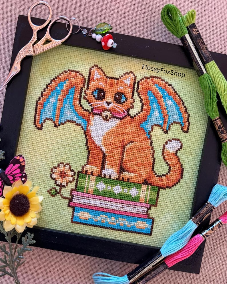 May include: Cross-stitch art of an orange cat with blue and orange wings, sitting on a stack of books. The artwork is framed in black and surrounded by embroidery floss, scissors, and a charm. The text "FlossyFoxShop" is visible.