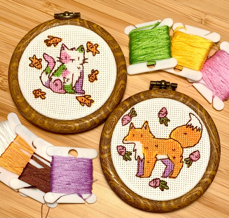 May include: Two embroidery hoops with cross-stitch designs. The left hoop features a pink and green cat with a white face and yellow fish. The right hoop features an orange fox with a white tail and purple legs. Both hoops have a wooden frame.
