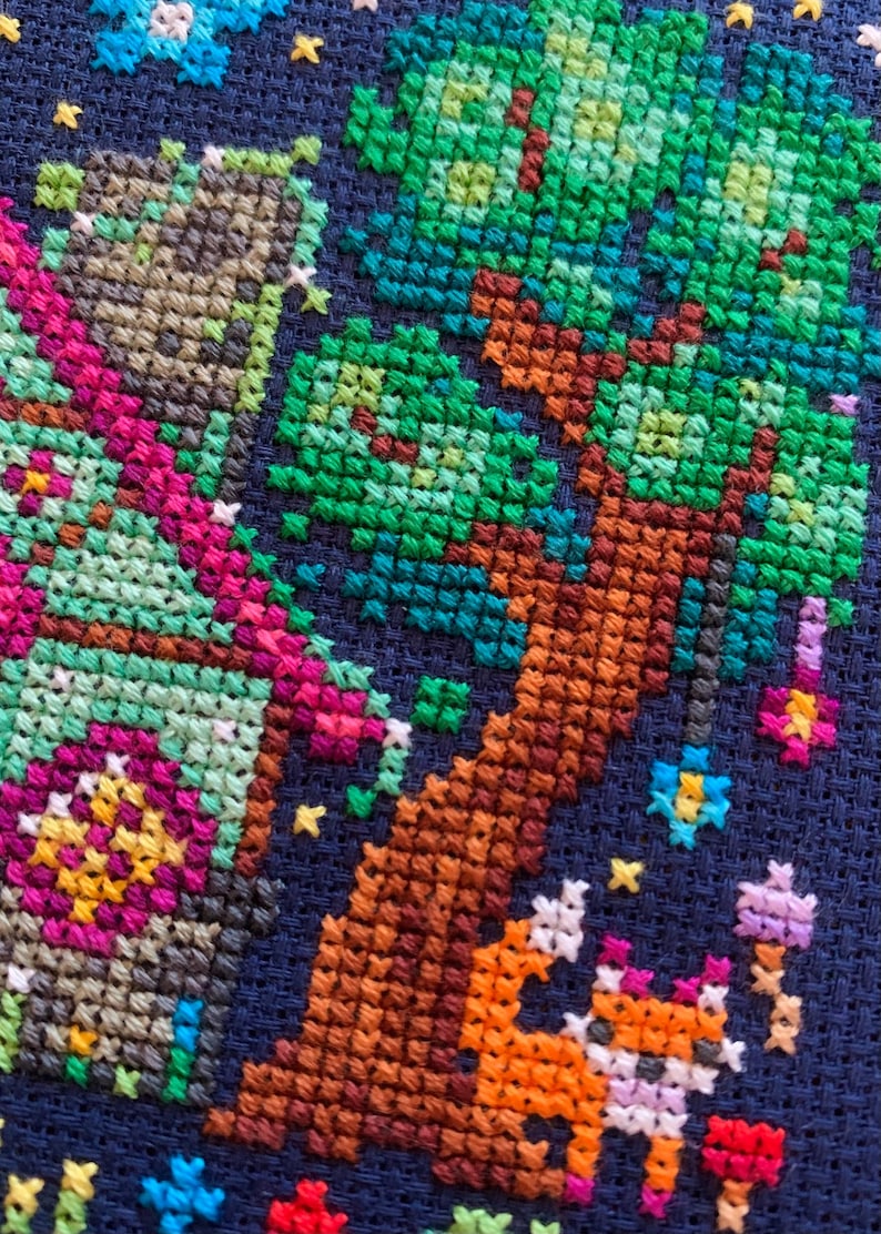 May include: Cross-stitch embroidery on dark blue fabric. The design features a tree with green leaves and a brown trunk, a fox, and floral and star accents. The colour palette includes shades of green, brown, orange, pink, and blue.