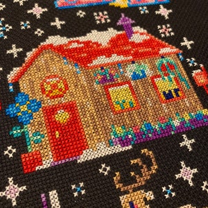May include: A cross-stitch pattern of a house with a red door, a reindeer, and a unicorn. The house is decorated with lights and a wreath. The background is a dark blue with snowflakes.