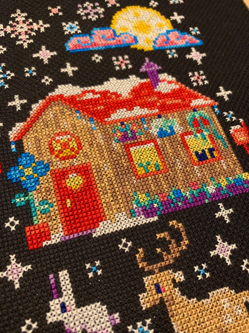 May include: A cross-stitch pattern of a house with a red door, a reindeer, and a unicorn. The house is decorated with lights and a wreath. The background is a dark blue with snowflakes.