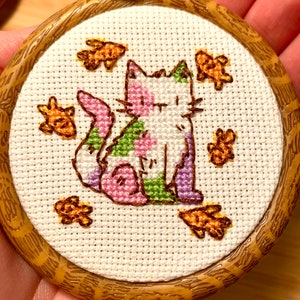 May include: A white cat with pink, green, and purple spots is surrounded by orange fish in a wooden embroidery hoop.