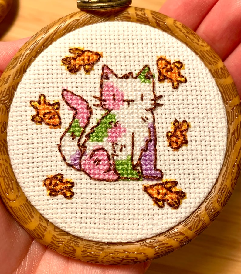 May include: A white cat with pink, green, and purple spots is surrounded by orange fish in a wooden embroidery hoop.