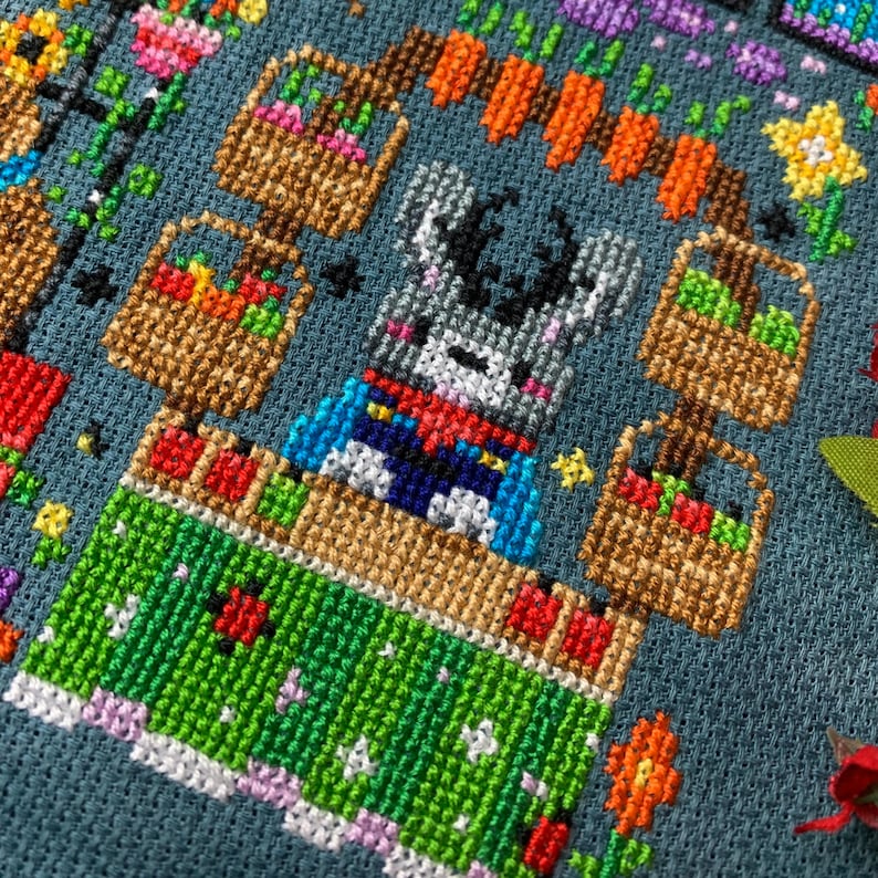 May include: Cross-stitch pattern of a gray bunny rabbit wearing a blue and red cape, standing in front of a stand with baskets of colorful fruit and vegetables. The stand is decorated with green leaves and flowers.