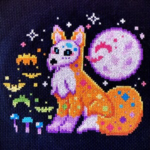 May include: A cross-stitch embroidery of a purple and orange fox with a pink moon behind it. The fox is surrounded by stars and other small details.
