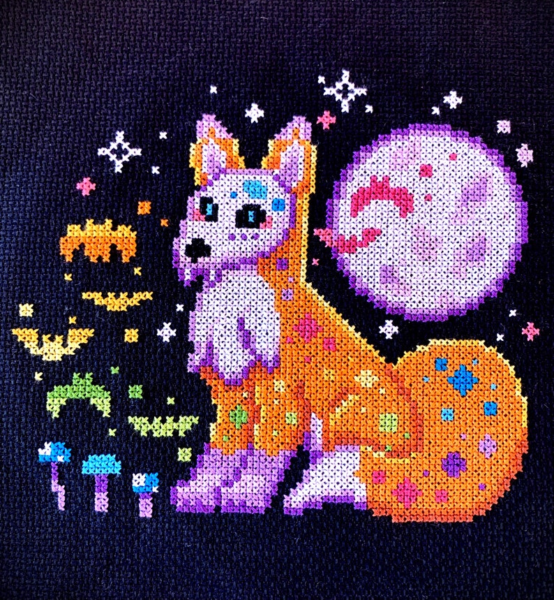 May include: A cross-stitch embroidery of a purple and orange fox with a pink moon behind it. The fox is surrounded by stars and other small details.